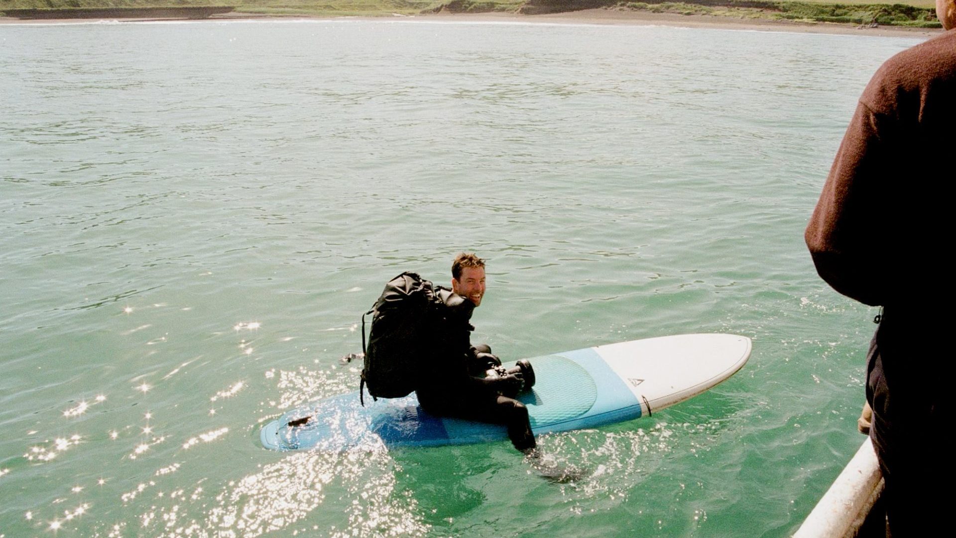Man wearing backpack full of camera gear sits on surf board in the water