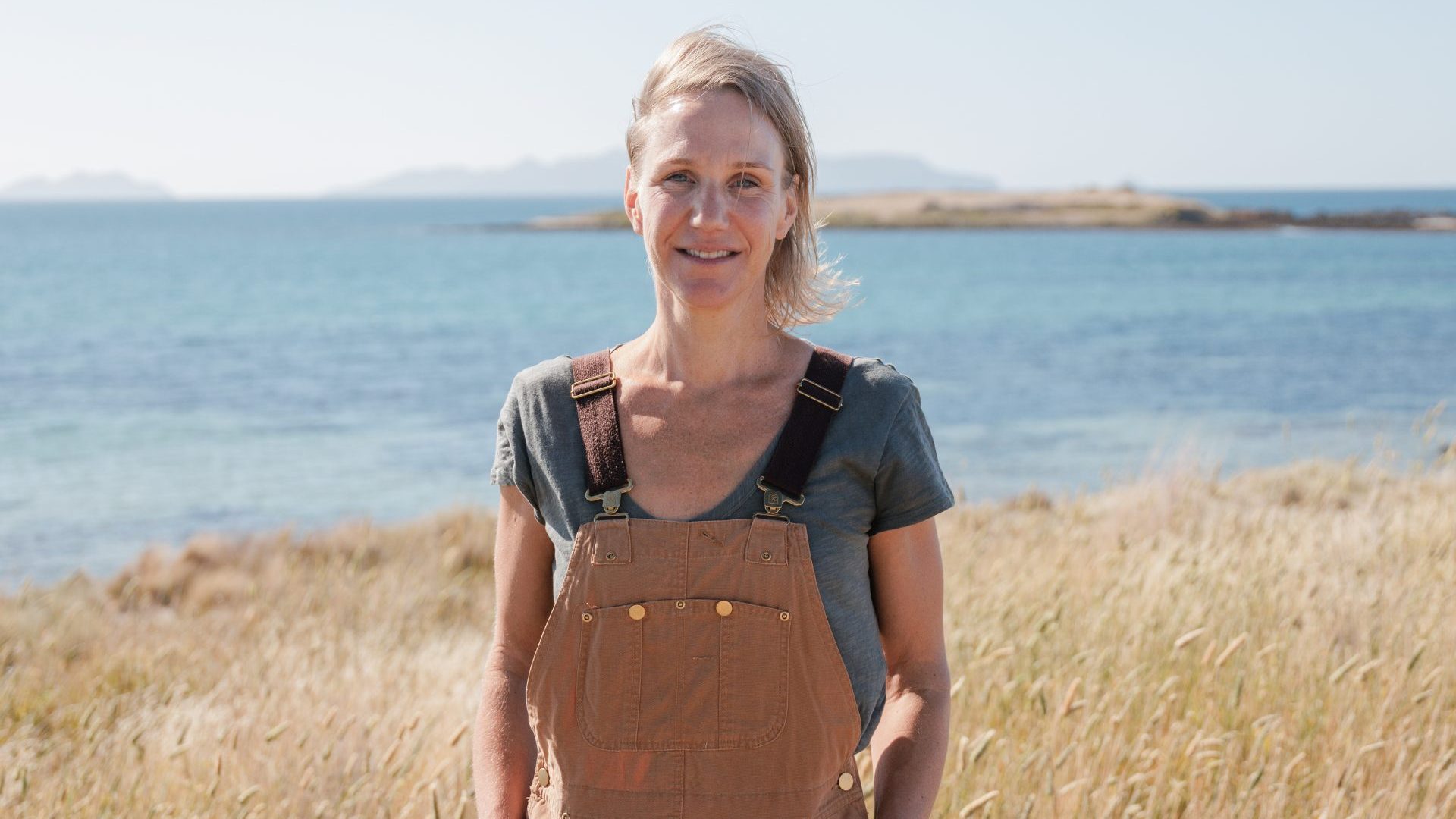 A woman in brown dungarees smiles to camera with the sea in the background.
