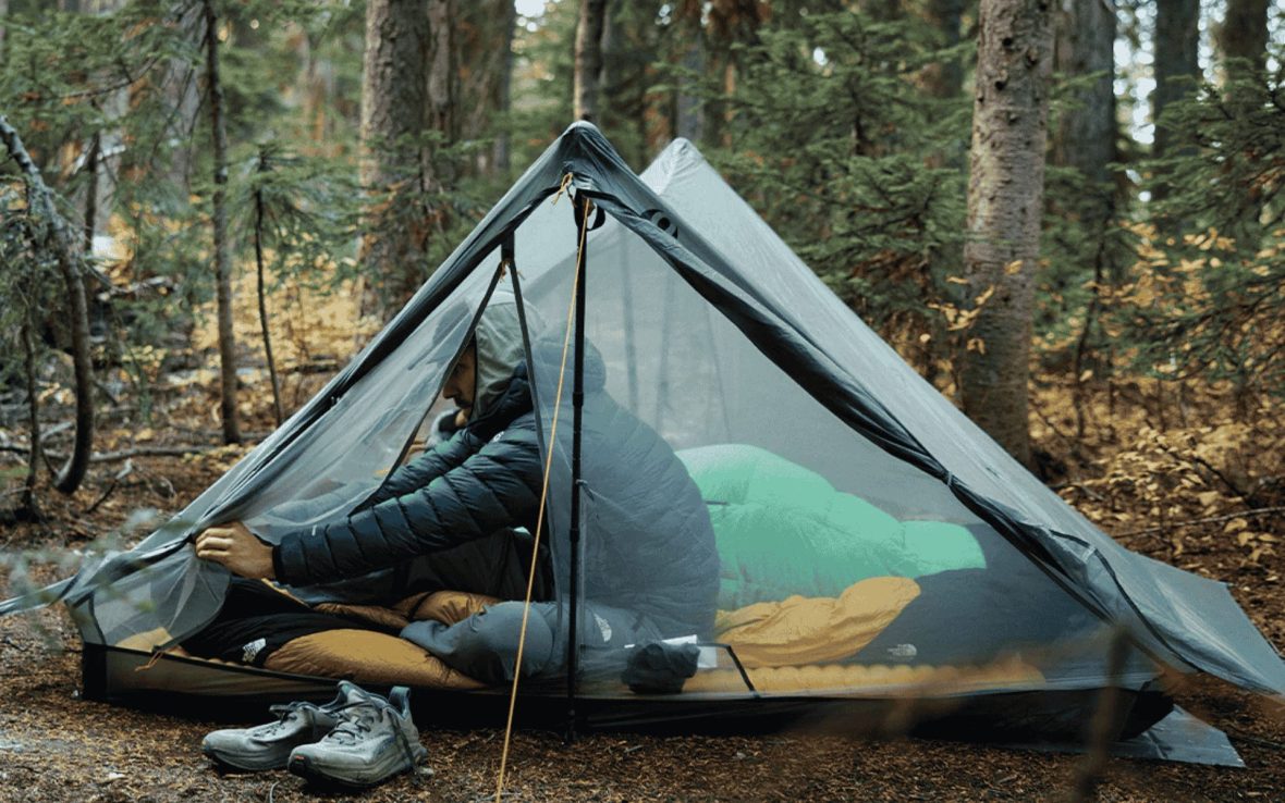 A camper reaches out from inside a triangular tent with fully-mesh doors. The forest is in the background.