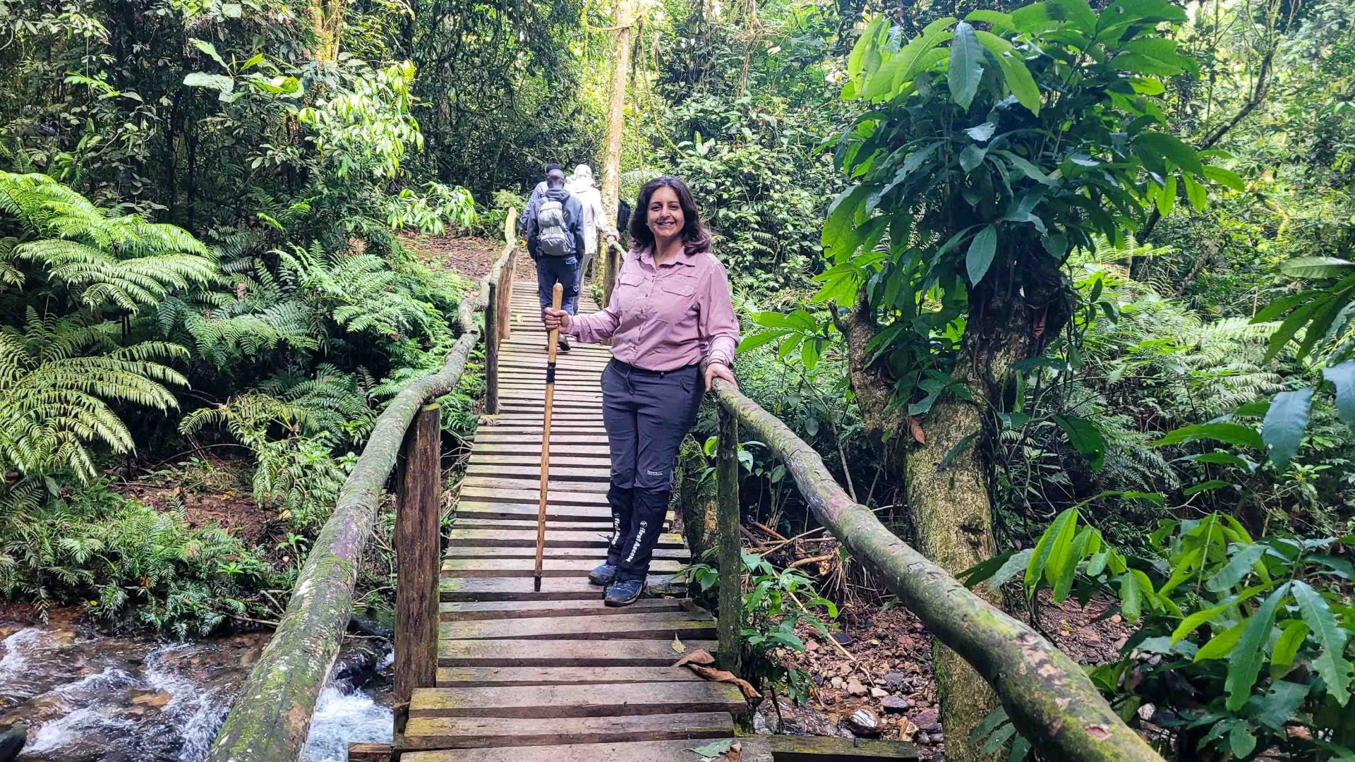 A woman leans on the railing of a wooden bridge in a dense forest.