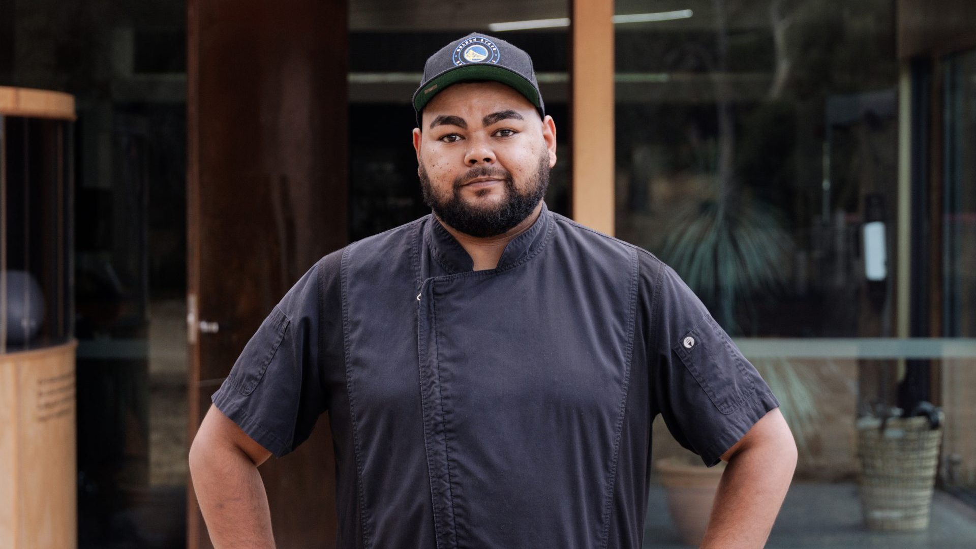 A man (chef Braydon Gower) with a beard in a dark cap and shirt looks into the camera.