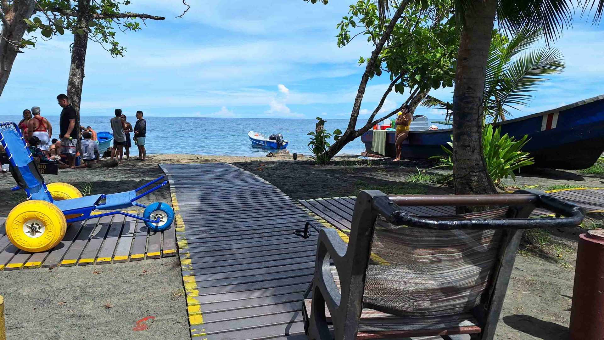 A wooden accessible boardwalk leads toward a beach with trees and boats. A blue and yellow beach wheelchair is off to the side of the boardwalk.
