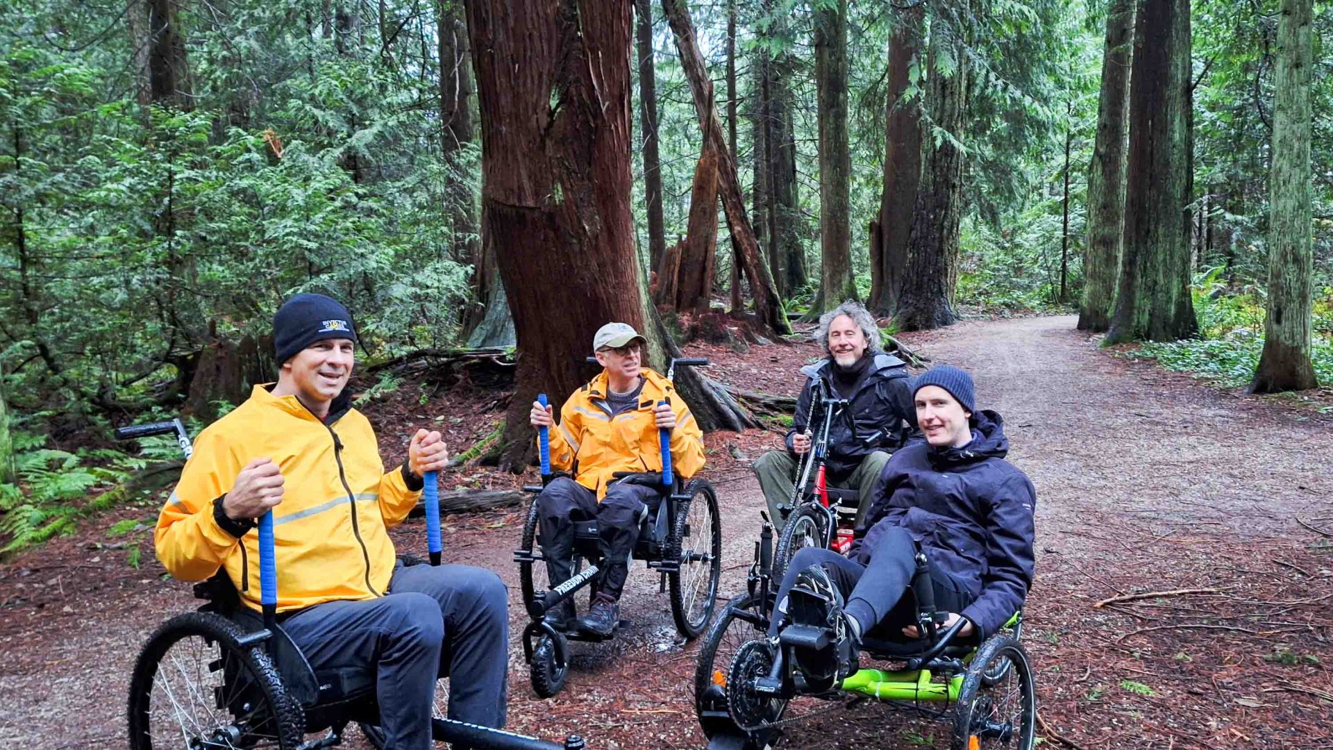 Four men in wheelchairs smile for a group portrait in a forest clearing. Two wear orange jackets and two wear black jackets.