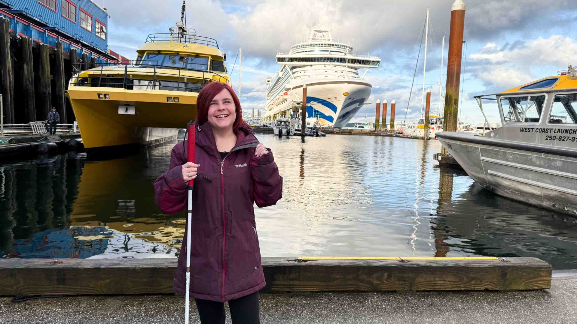 A woman with a purple coat and white cane stands in front of a harbour with boats in it.