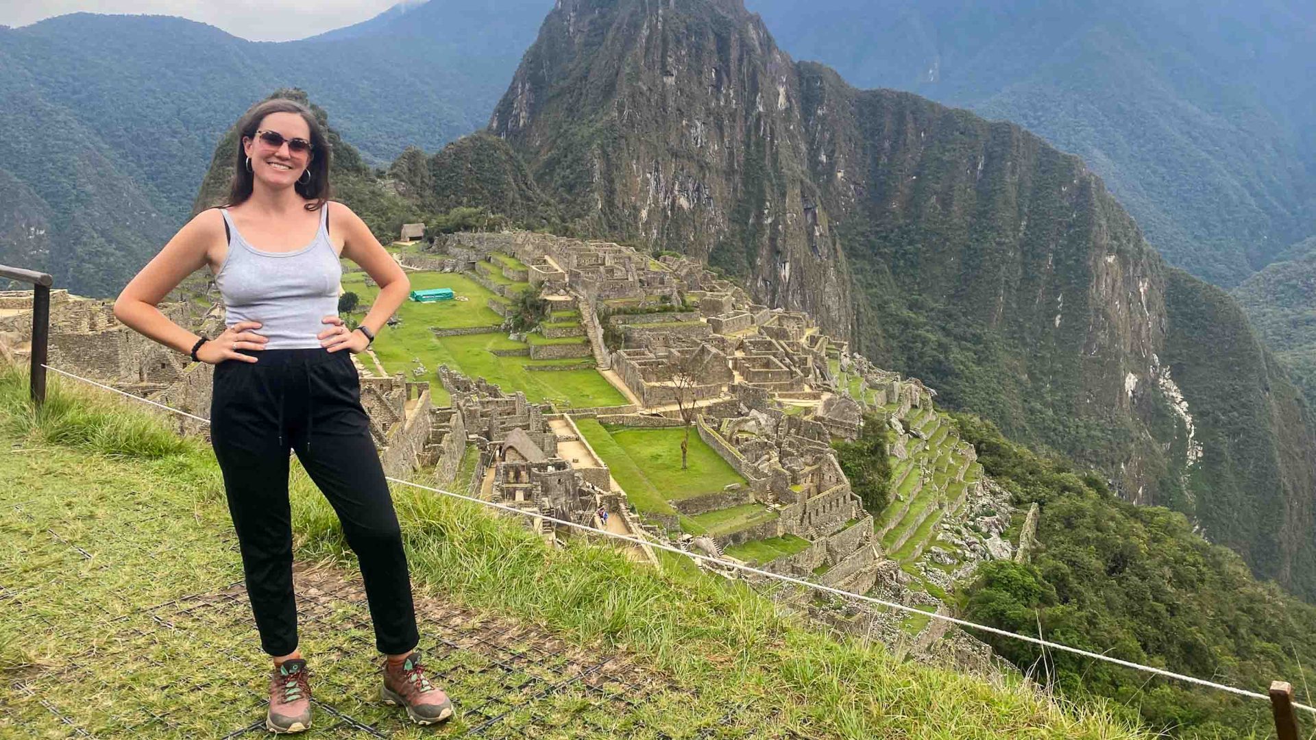 A woman in black leggings and grey top smiles as she stands in front of Machu Picchu.