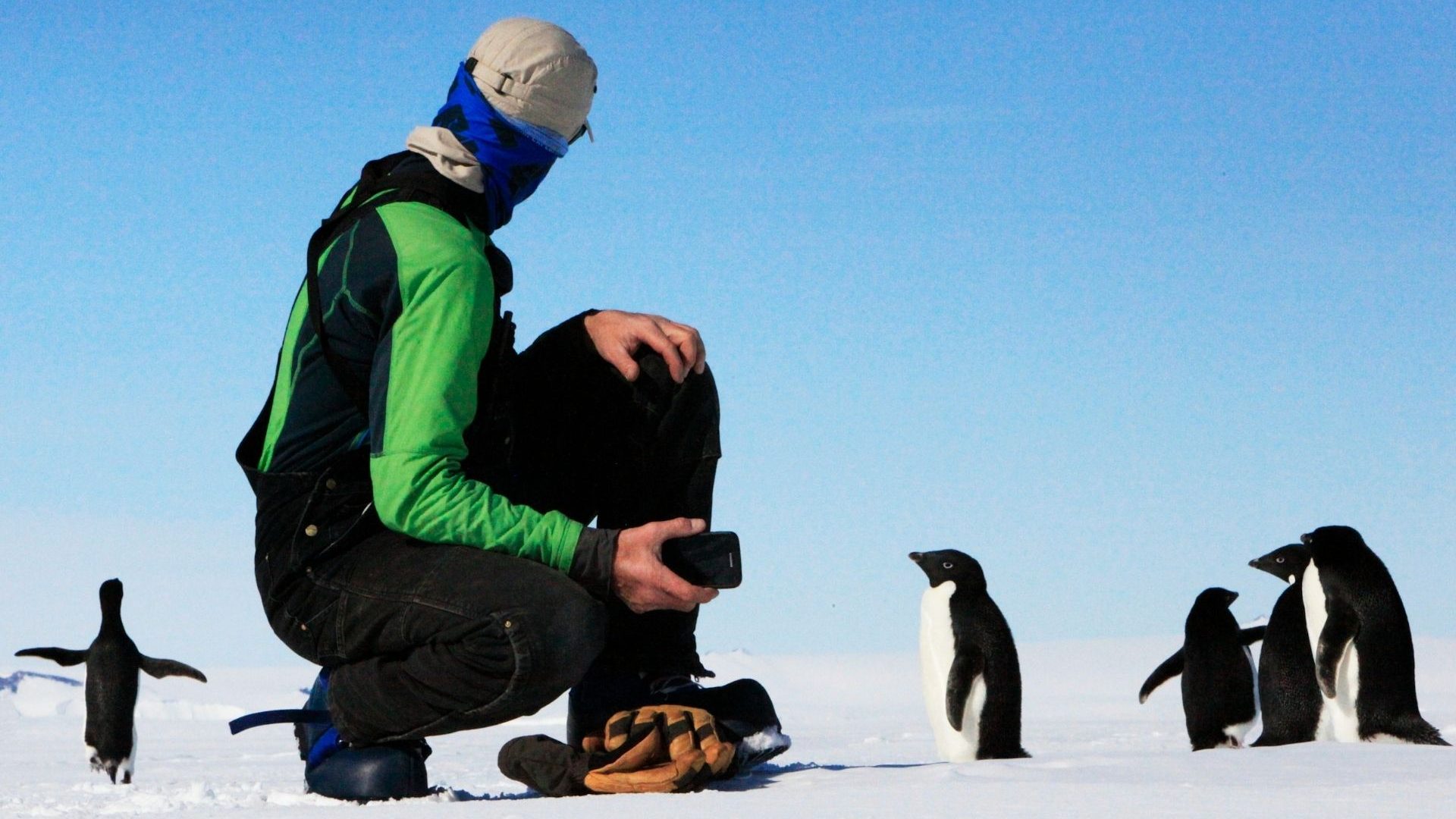 A close-up of a tourist crouching on the ice with a penguin a couple of feet away.