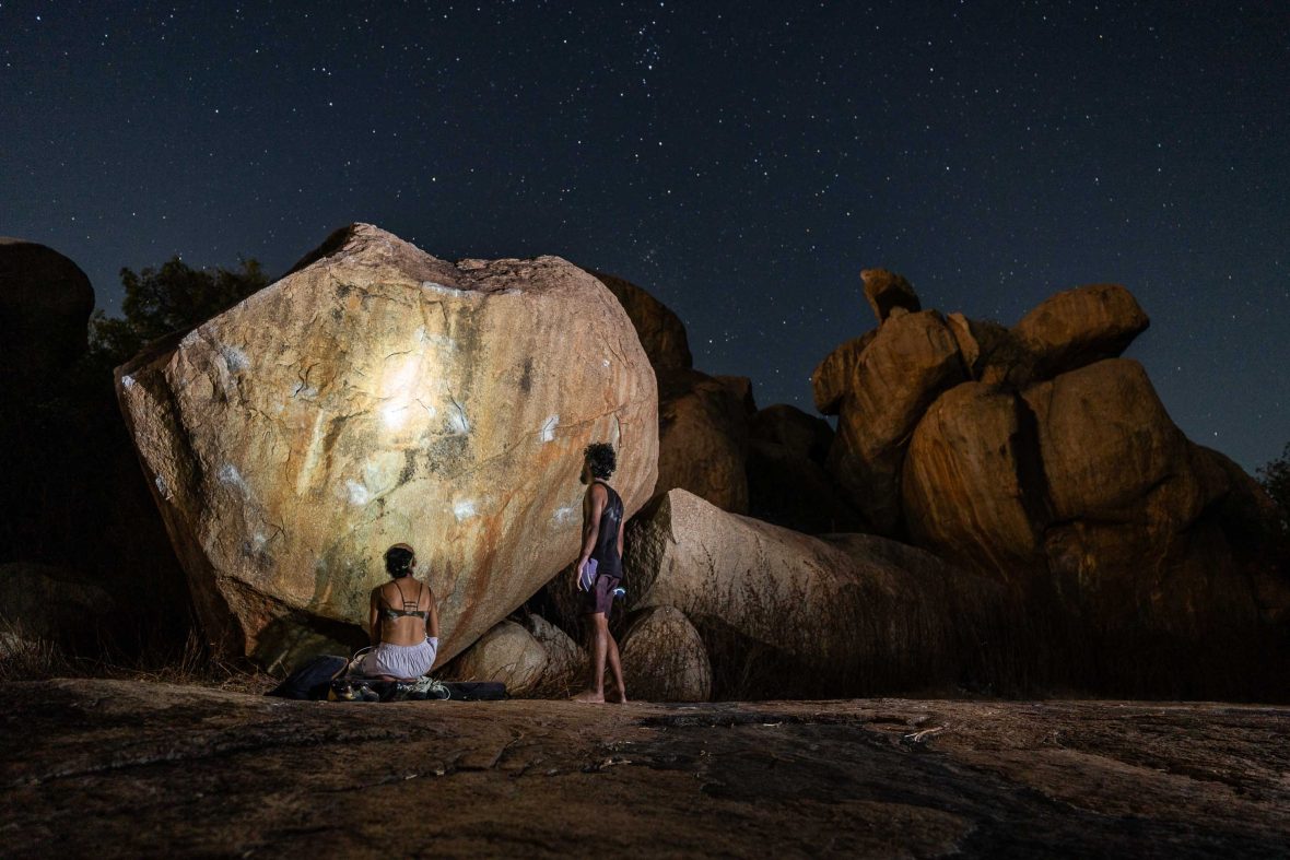 Two people look at a boulder at night and appear to be having a conversation.
