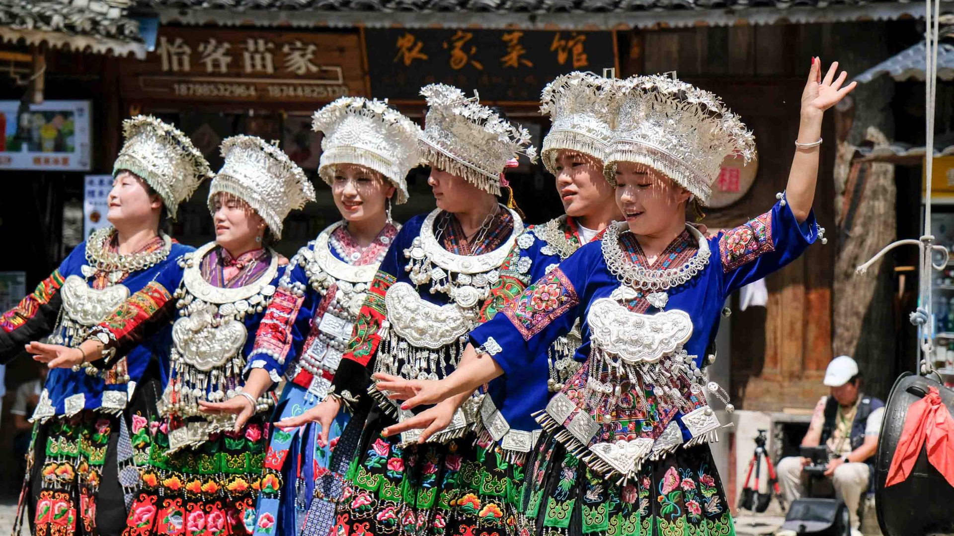Villagers in traditional blue clothing and silver head dresses do a performance in a town square.