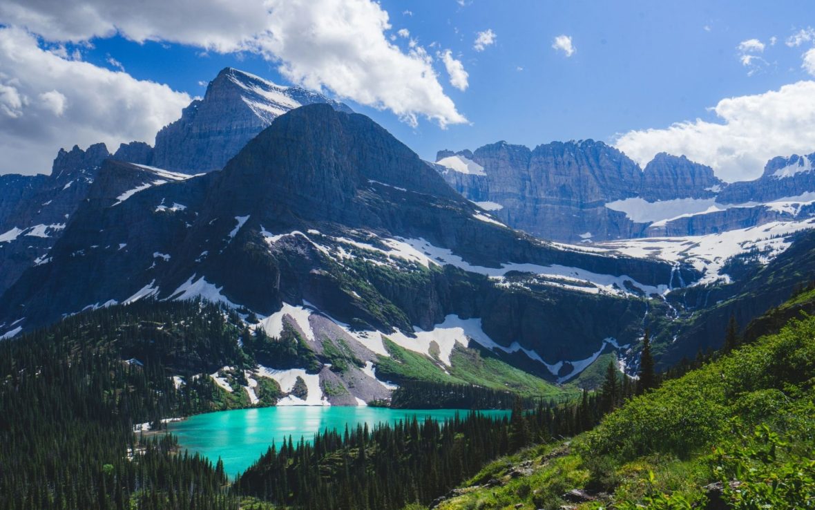 A view of mountains in Glacier National Park on a partly-cloudy day, with blue skies and a turquoise lake.