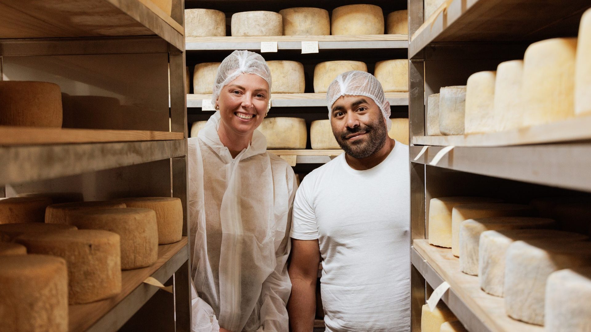 A man and woman smile to camera in a cheese room.