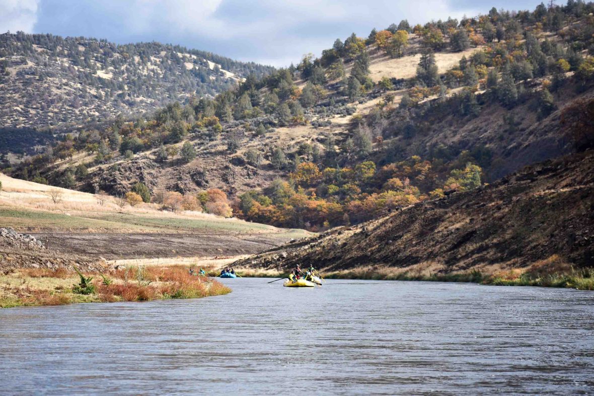 A yellow white water raft goes down a river with hills behind it with green trees on them.