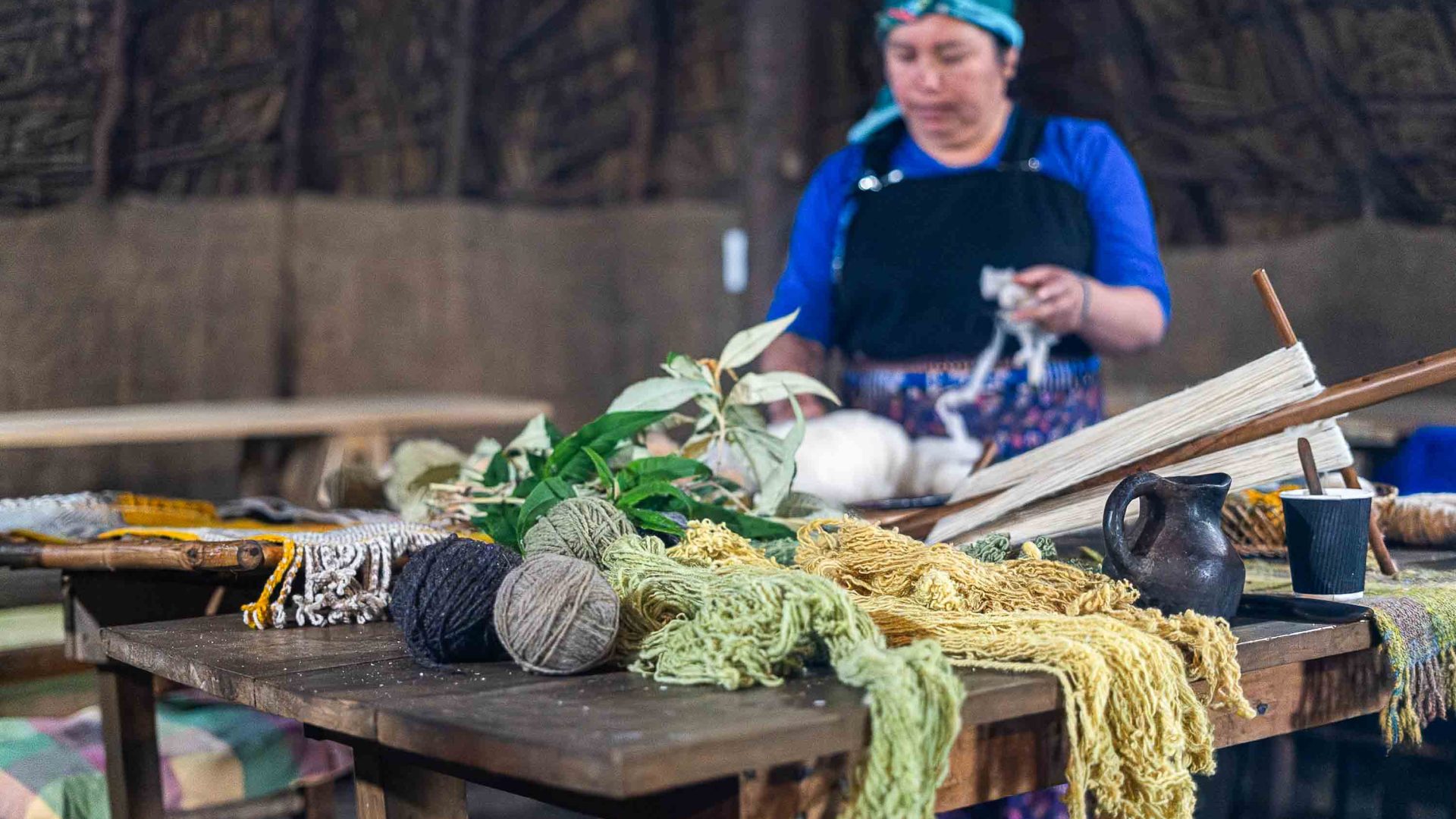 A woman weaves. There is a collection of wool on the table in front of her.