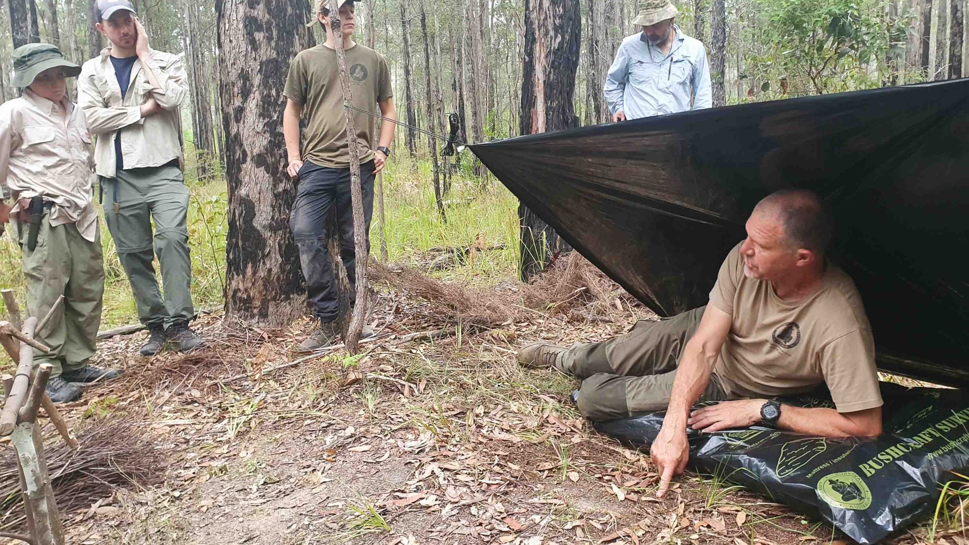 A man lies underneath a tarp that is on a 45 degree angle and talks to a group.