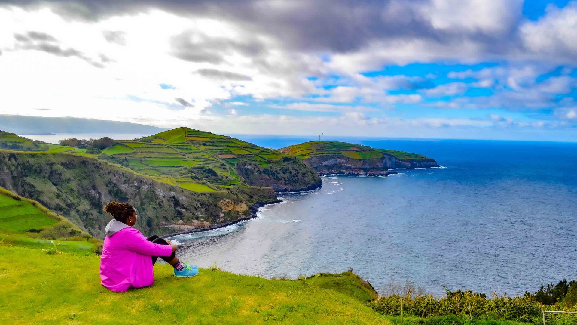 A woman with pink looks out over the water and hills.
