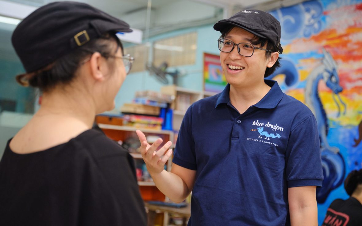 Man wearing blue shirt embroidered with 'Blue Dragon Childen's Foundation' smiles in conversation with a colleague.