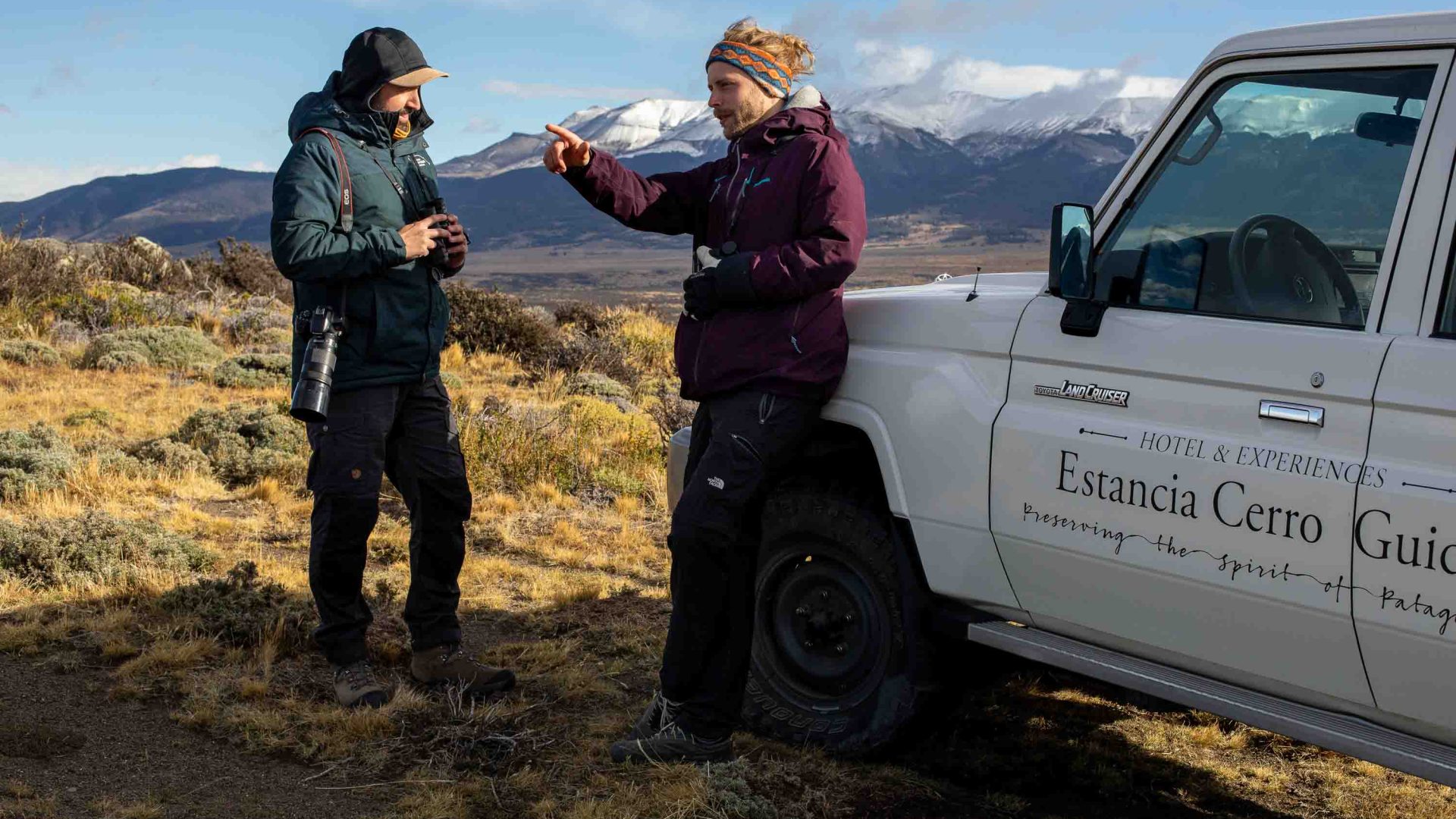 Two men stand by a vehicle and one of them points to the distance.