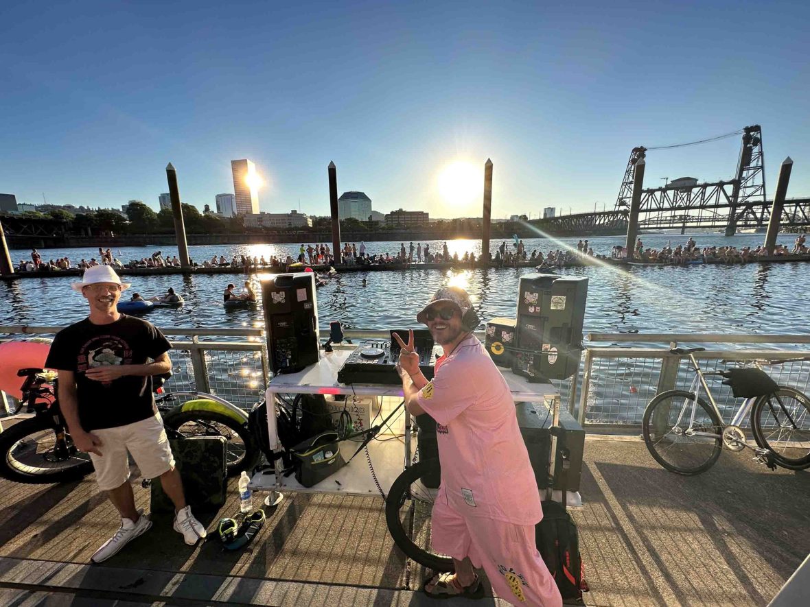 A DJ smiles to camera as he performs in front of people relaxing on the edge of a river.