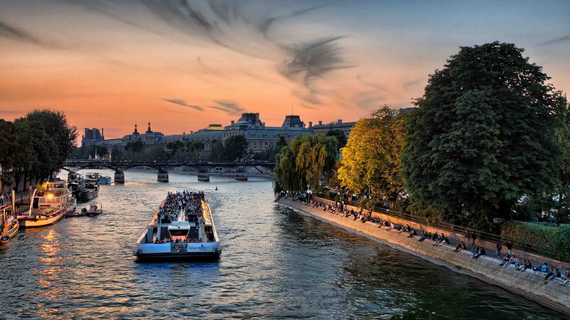 People sit on the edge of a river while a boat passes them.