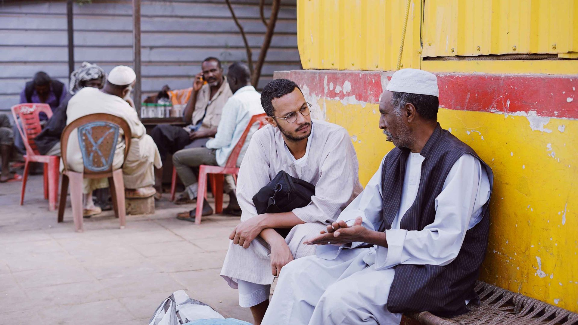 Two men sit and talk against a yellow wall in a merket.