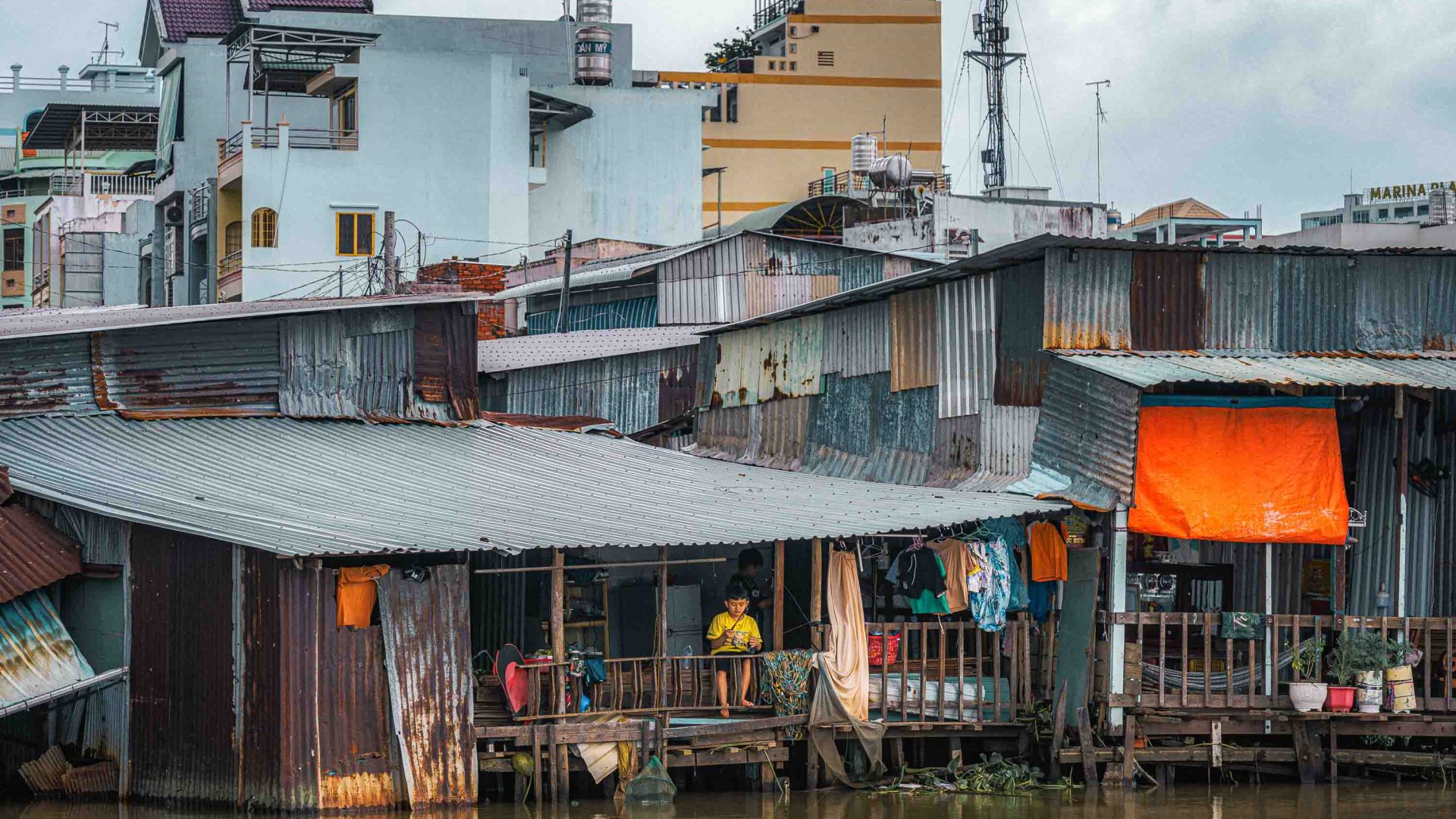 A young buy sits on the porch of his river house.